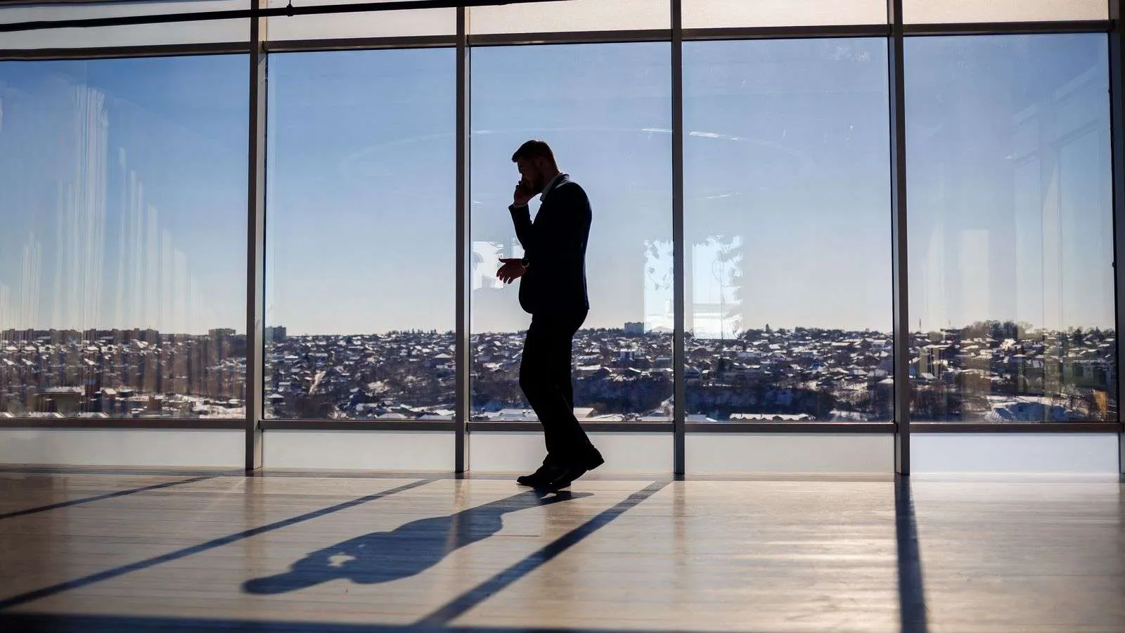 The silhouette of a man on a phone call by tall office windows overlooking a city